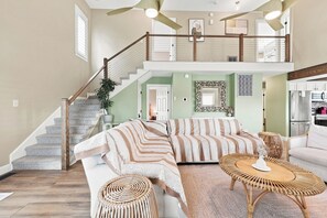 A spacious living area with a staircase leading to an upper level, a sofa set, a wicker coffee table, and a kitchen area. - Palm Cove | Luxury Channel Home + Coastal Escape (North Myrtle Beach)