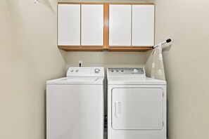 Een wasserette met een voorlader wasmachine en droger, houten kastjes erboven, en een hangend strijkijzer aan de rechterkant.