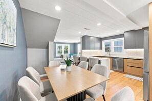 A modern kitchen with a built-in dishwasher, a dining area with a wooden table and chairs, and a view of the outdoors through large windows. - Elvi's Beach House | Spacious 6BR Duplex (North Myrtle Beach)
