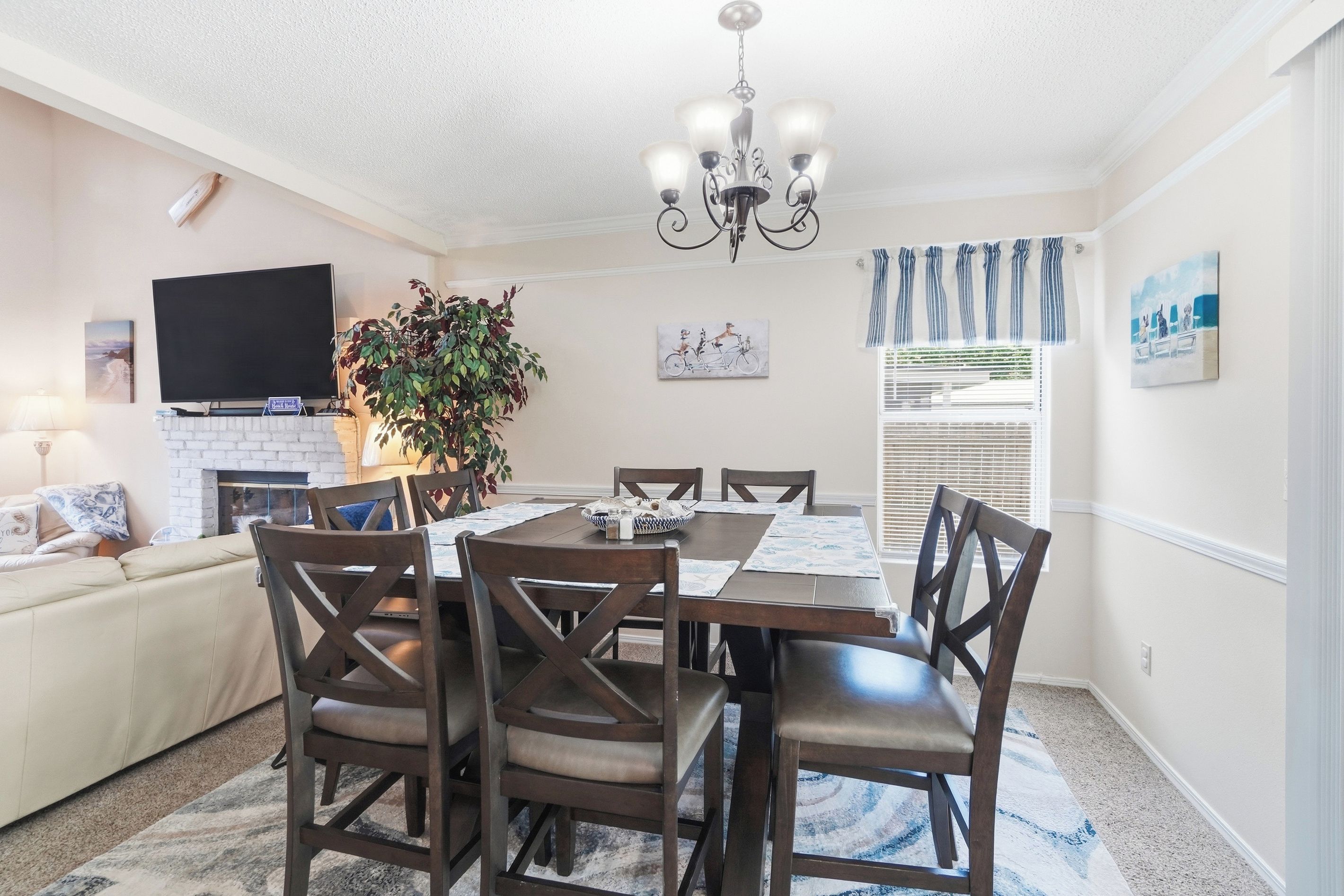 A dining area with a table, chairs, and a chandelier. There is a television on the wall and a plant in the corner.