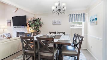 A dining area with a table, chairs, and a chandelier. There is a television on the wall and a plant in the corner.