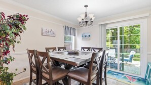Un coin repas avec une table dressée pour quatre personnes, entourée de chaises, d’un lustre et offrant une vue sur une terrasse extérieure, accessible par une porte vitrée.