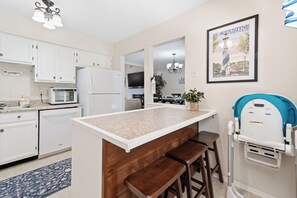 A kitchen with white cabinets, a microwave, and a refrigerator. There is a dining area with a table and chairs, and a poster on the wall. - Family 4BR Home | Fenced Yard | 5 Min to Beach (Jacksonville)