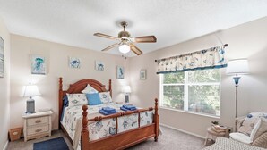 Une chambre à coucher avec un lit en bois, deux tables de chevet avec des lampes, un ventilateur de plafond, une fenêtre avec des stores et des œuvres d’art accrochées aux murs.