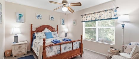 A bedroom with a wooden bed, two bedside tables with lamps, a ceiling fan, a window with blinds, and artwork on the walls.