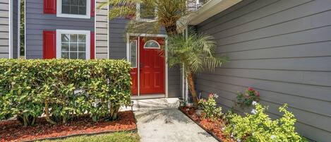A residential building with a red door, a pathway leading to the entrance, and a variety of plants and shrubs.