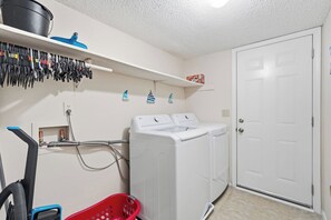 A laundry room with a washing machine, dryer, and a hose. There are shelves with tools and a red laundry basket. - Family 4BR Home | Fenced Yard | 5 Min to Beach (Jacksonville)