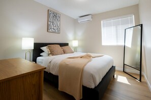 A bedroom with a bed, bedside lamps, a wooden nightstand, a wall-mounted air conditioner, and a window with blinds. - Cozy Casa | Contemporary 2BR Home · La Mesa (La Mesa)