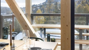 Zona pranzo con tavolo apparecchiato per due, vista su un paesaggio innevato attraverso ampie finestre e un'uscita su un balcone in legno.
