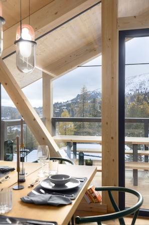 A dining area with a table set for two, a view of a snowy landscape through large windows, and a wooden deck outside. - 03_NTH029_Premium Chalet mit Sauna und Hot Tub by Interhome (Turrach)