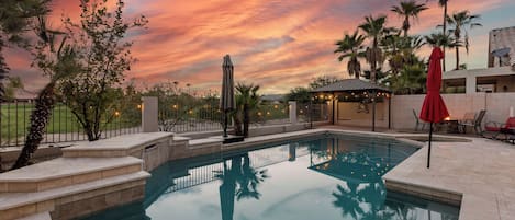 Una piscina con vista al atardecer, rodeada de palmeras y una zona de estar cubierta.