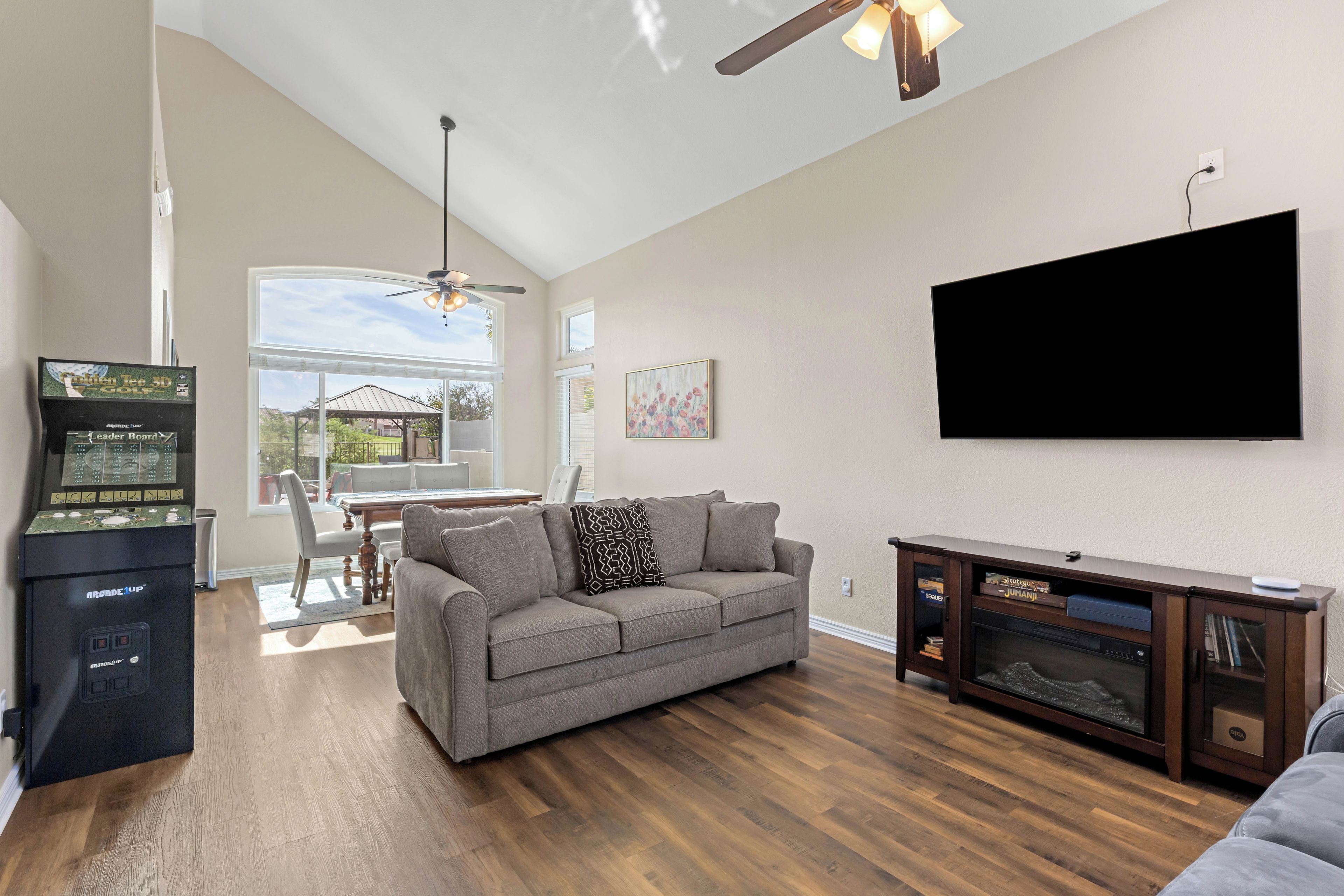 A living room with a grey sofa, wooden flooring, a ceiling fan, a mounted TV, and a gaming console.