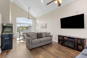 Ein Wohnzimmer mit grauem Sofa, Holzboden, Deckenventilator, montiertem Fernseher und Spielkonsole.