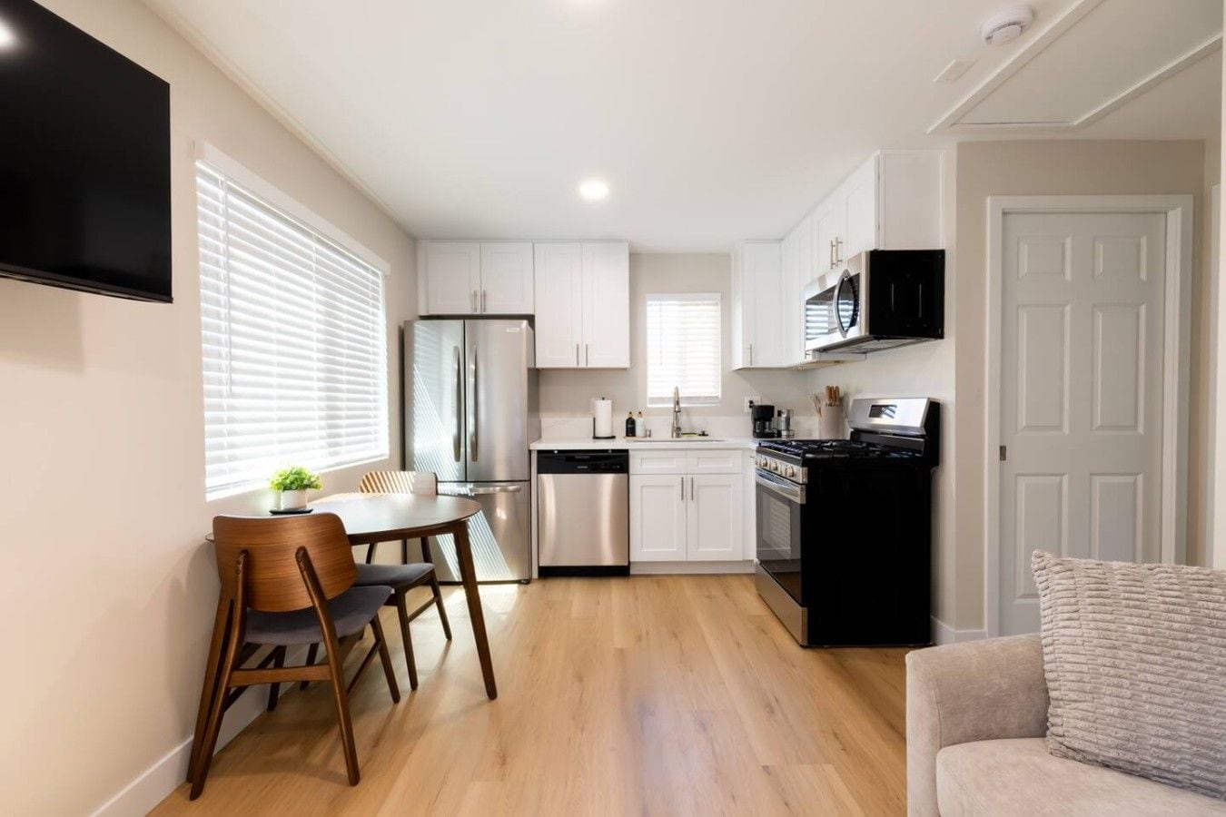 A compact kitchen with white cabinets, a stainless steel refrigerator, a black stove, and a dining area with a wooden table and chairs.