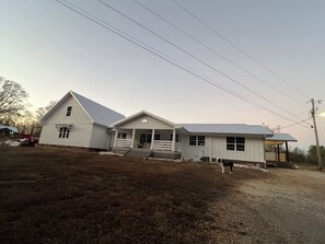 Uma casa branca de dois andares com varanda e área coberta, cercada por um caminho de cascalho e gramado.
