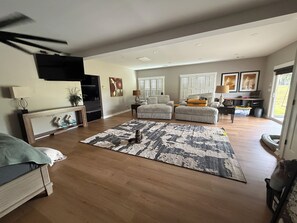 Uma sala de estar ampla, com um tapete grande, um sofá, uma poltrona e uma televisão presa na parede.