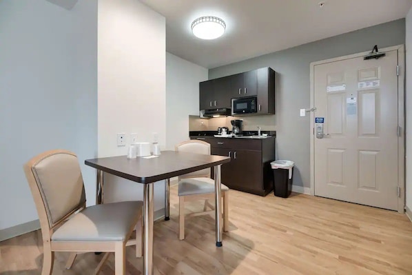 Cocina compacta con microondas, armarios y un cesto de basura. Hay una mesa de comedor con sillas y una lámpara en el techo.
