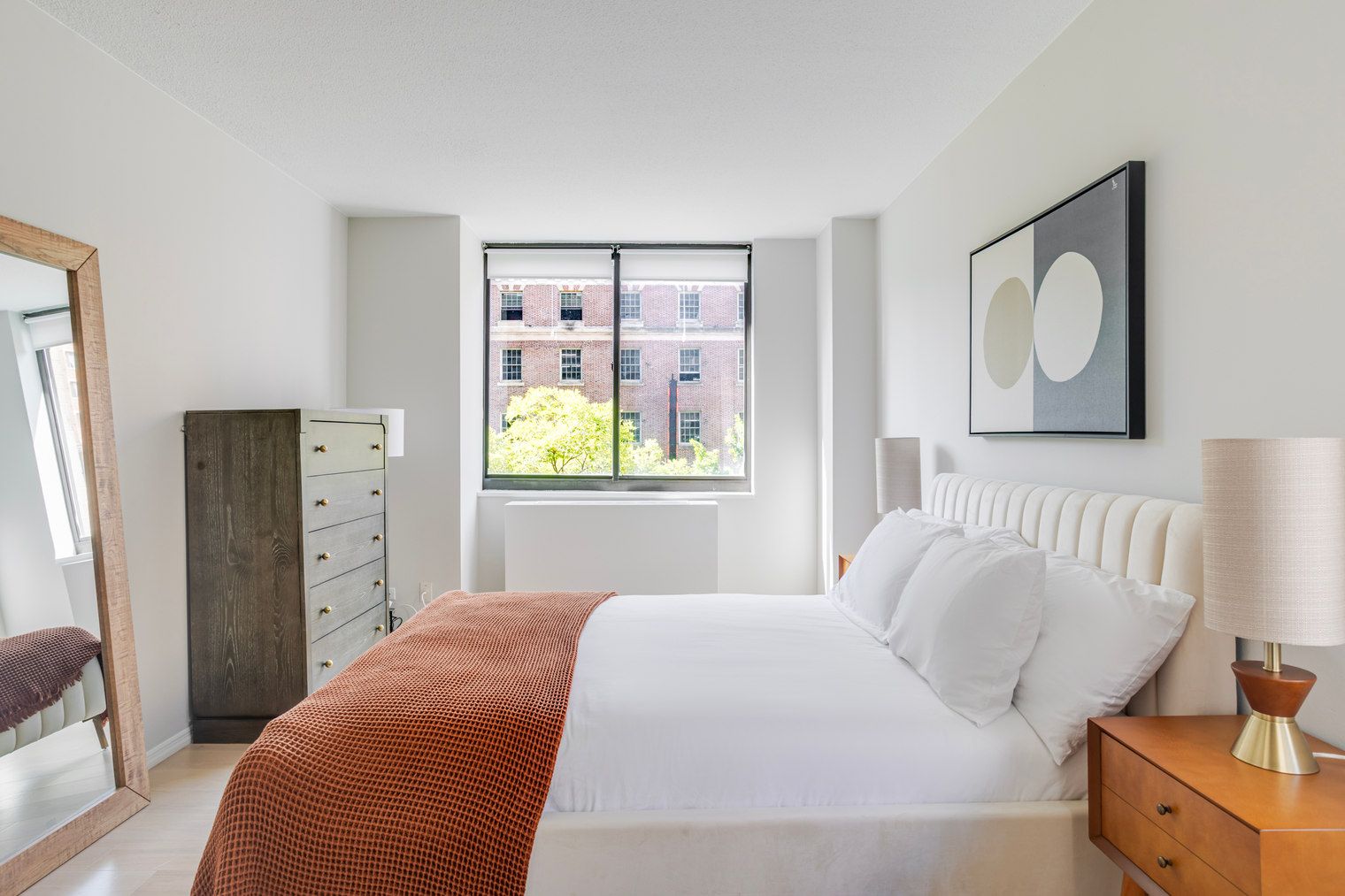A modern bedroom with a bed, a dresser, a window with a view of buildings, and a lamp.