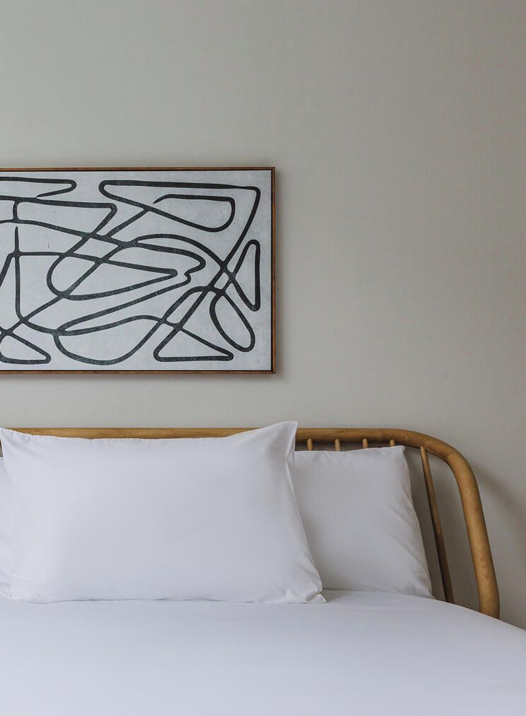Una cama con ropa blanca y cabecera de madera, una pared lisa y una obra de arte abstracta sobre la cama.