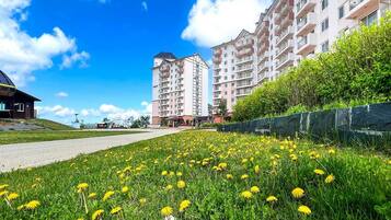 A residential building with multiple balconies, a paved road in the foreground, and a field of yellow flowers.