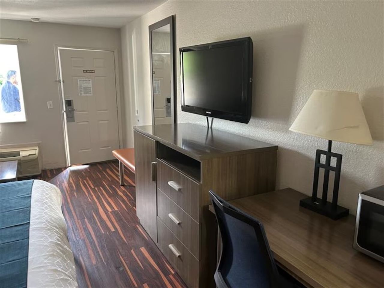 A hotel room with a bed, desk, chair, TV, lamp, and a door.