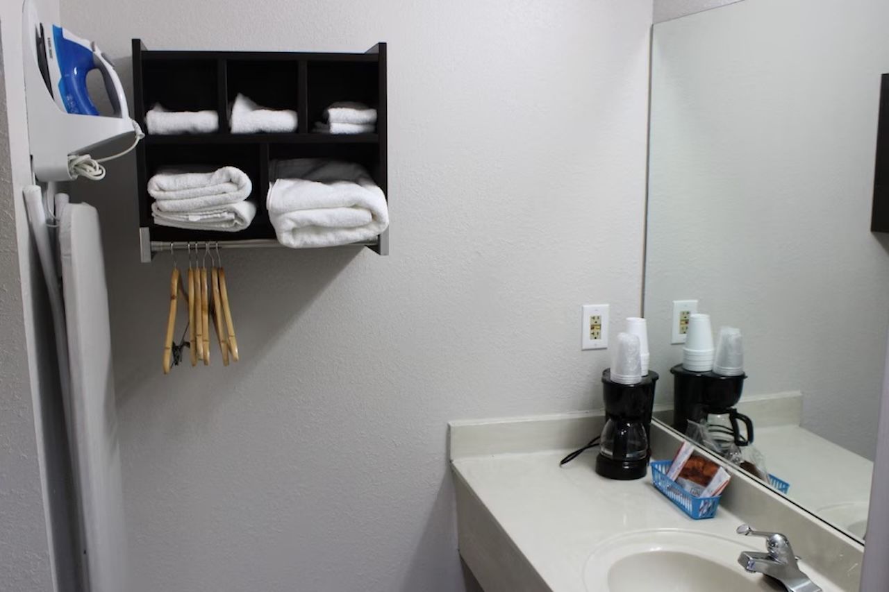 A bathroom with a white sink, a mirror, a towel rack with folded towels, a coffee maker, and a shelf with toiletries.