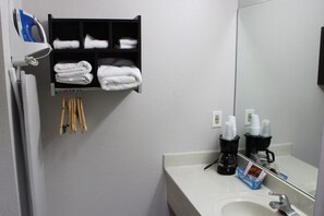 Un baño con lavamanos blanco, espejo, toallero con toallas dobladas, cafetera y repisa con artículos de aseo.