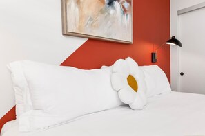 Una cama con ropa de cama blanca y un almohadón grande blanco con un centro amarillo, una lámpara de escritorio negra y un cuadro en la pared.