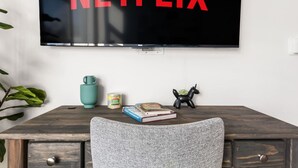 A room with a television mounted on the wall displaying 'NETFLIX' in red letters, a wooden desk with a chair, a small plant, a teal mug, a can of food, and a book. - The DTLA Haven, 1Mi to Dodger Stadium, Free Parking (Los Angeles)