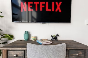 Habitación con una televisión en la pared mostrando 'NETFLIX' en letras rojas, un escritorio de madera con una silla, una planta pequeña, una taza azul verdosa, una lata de comida y un libro.