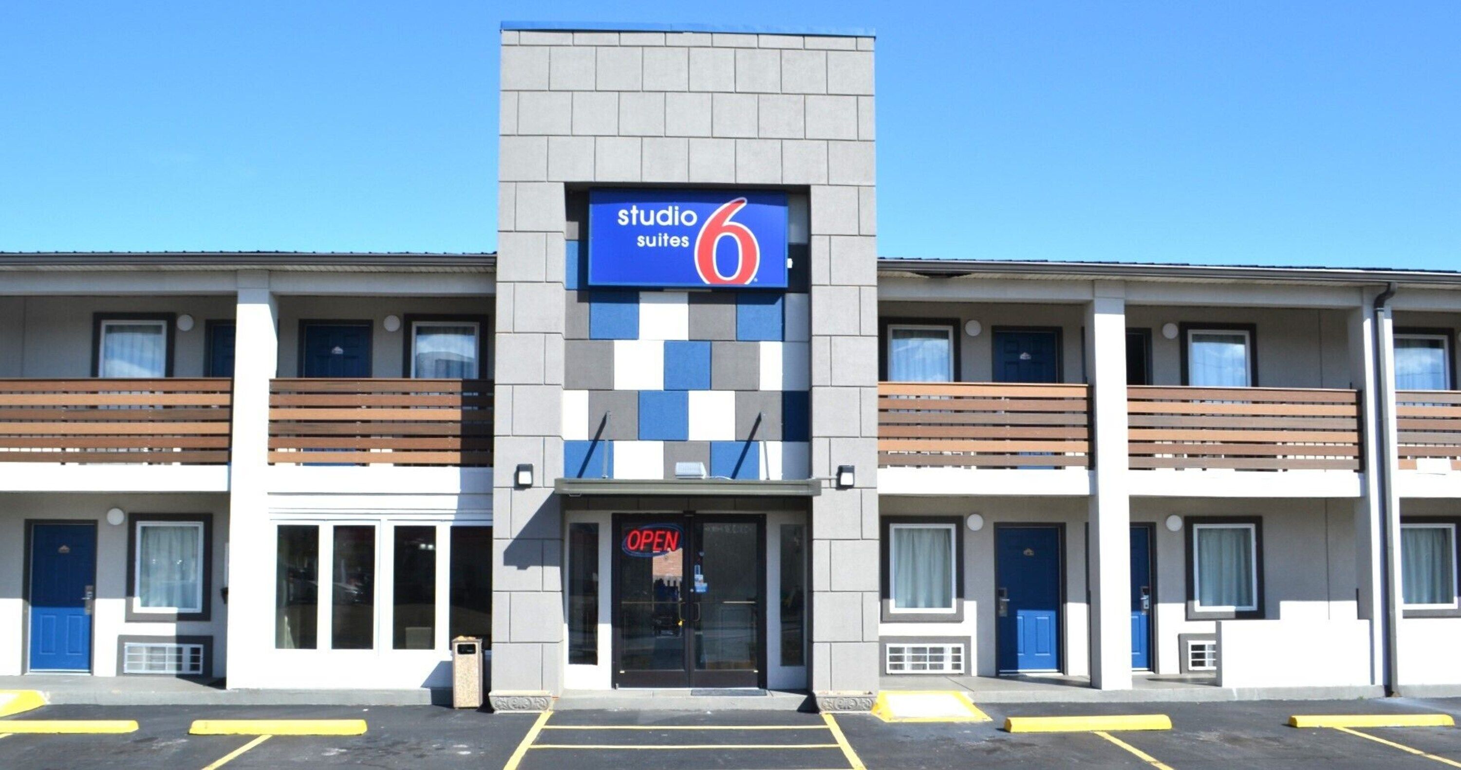 Un hôtel avec une enseigne indiquant « Studio 6 Suites », doté d’un design moderne, d’une entrée centrale, de plusieurs balcons et d’un parking avec des places délimitées.
