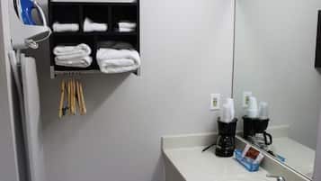A bathroom with a white sink, a mirror, a towel rack with folded towels, a coffee maker, and a shelf with toiletries.