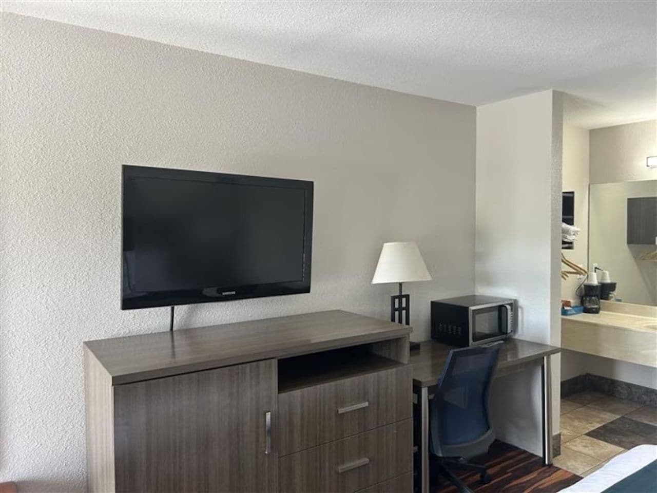 A hotel room with a flat-screen TV mounted on the wall, a desk with a lamp, a microwave, and a chair.
