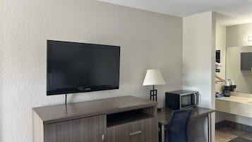 A hotel room with a flat-screen TV mounted on the wall, a desk with a lamp, a microwave, and a chair.
