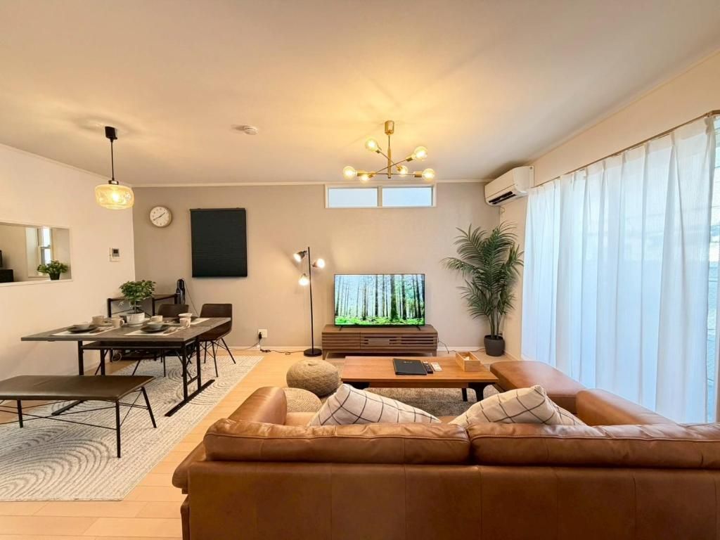 A modern living room with a leather sofa, a dining table, a TV, and a large window with curtains.