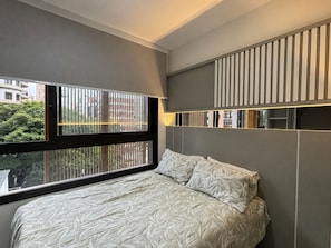 A modern bedroom with a large window, a bed with patterned bedding, and a wall-mounted light fixture. - Cozy 1-bedroom apartment with AC in lovely Moema neighbourhood in São Paulo (São Paulo)