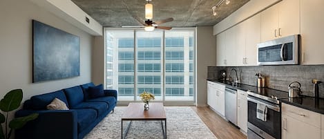 A modern kitchen with a microwave, stove, and cabinets, a blue sofa, a coffee table, and a large window with blinds.
