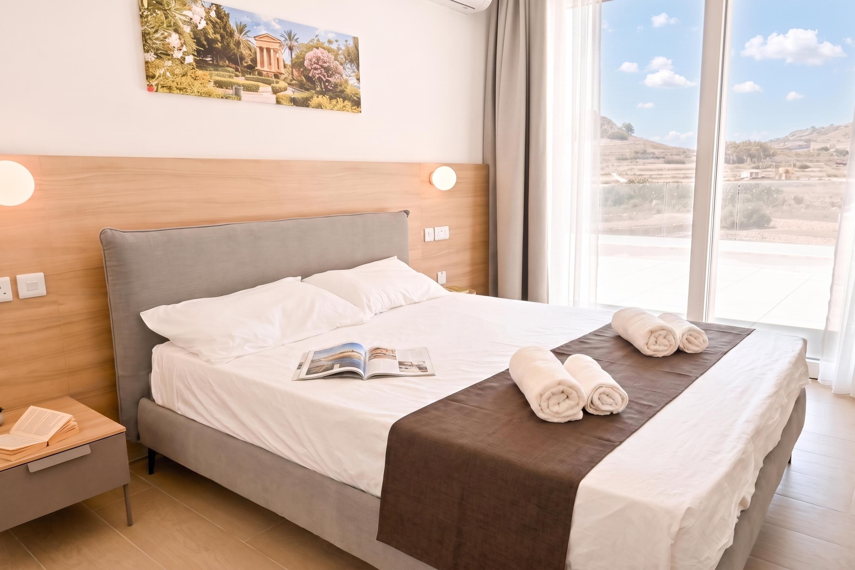 A hotel room with a large bed, a nightstand, a reading lamp, and a window with a view of the countryside.