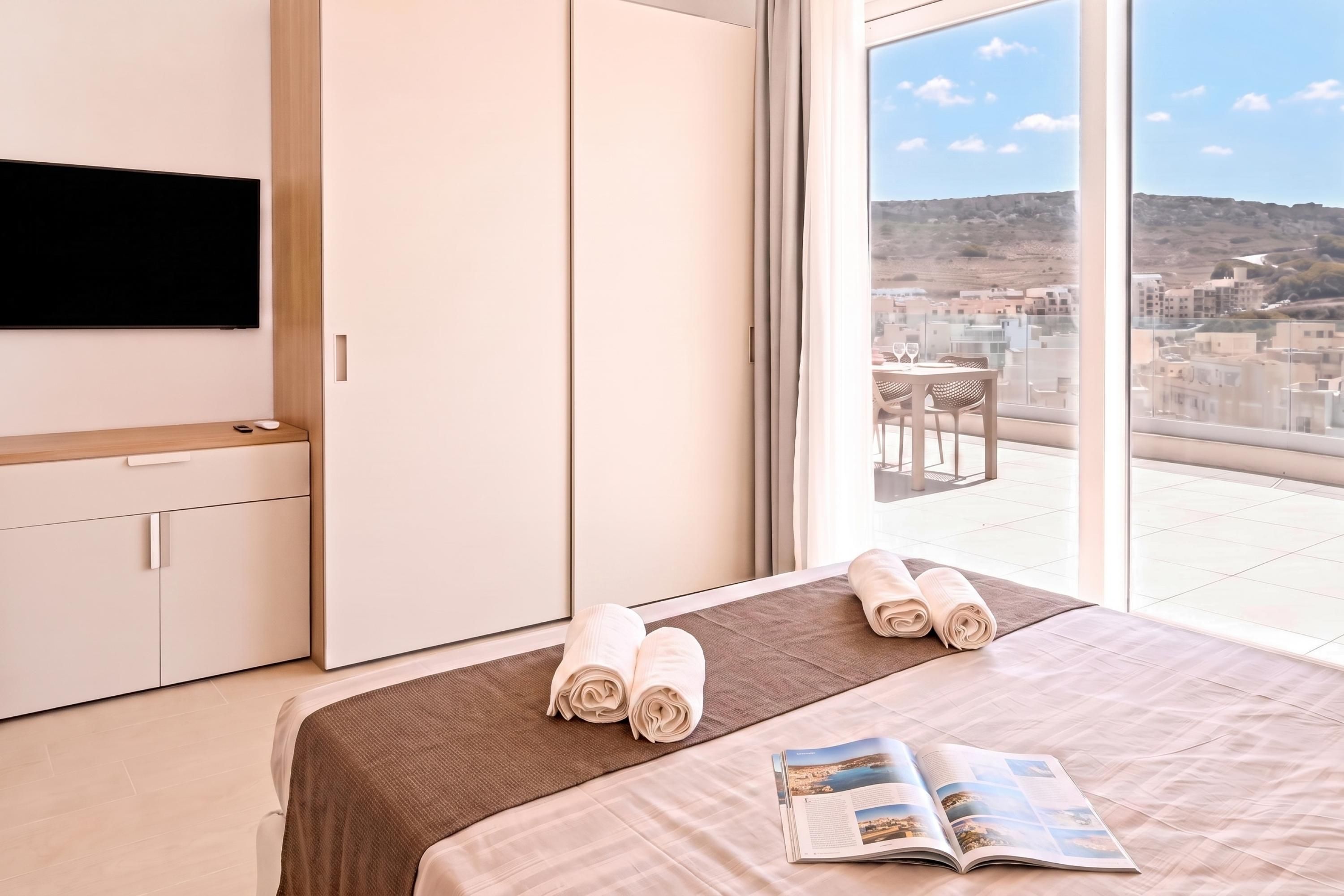 A hotel room with a bed, a flat-screen TV, a window with a city view, and a table with chairs.