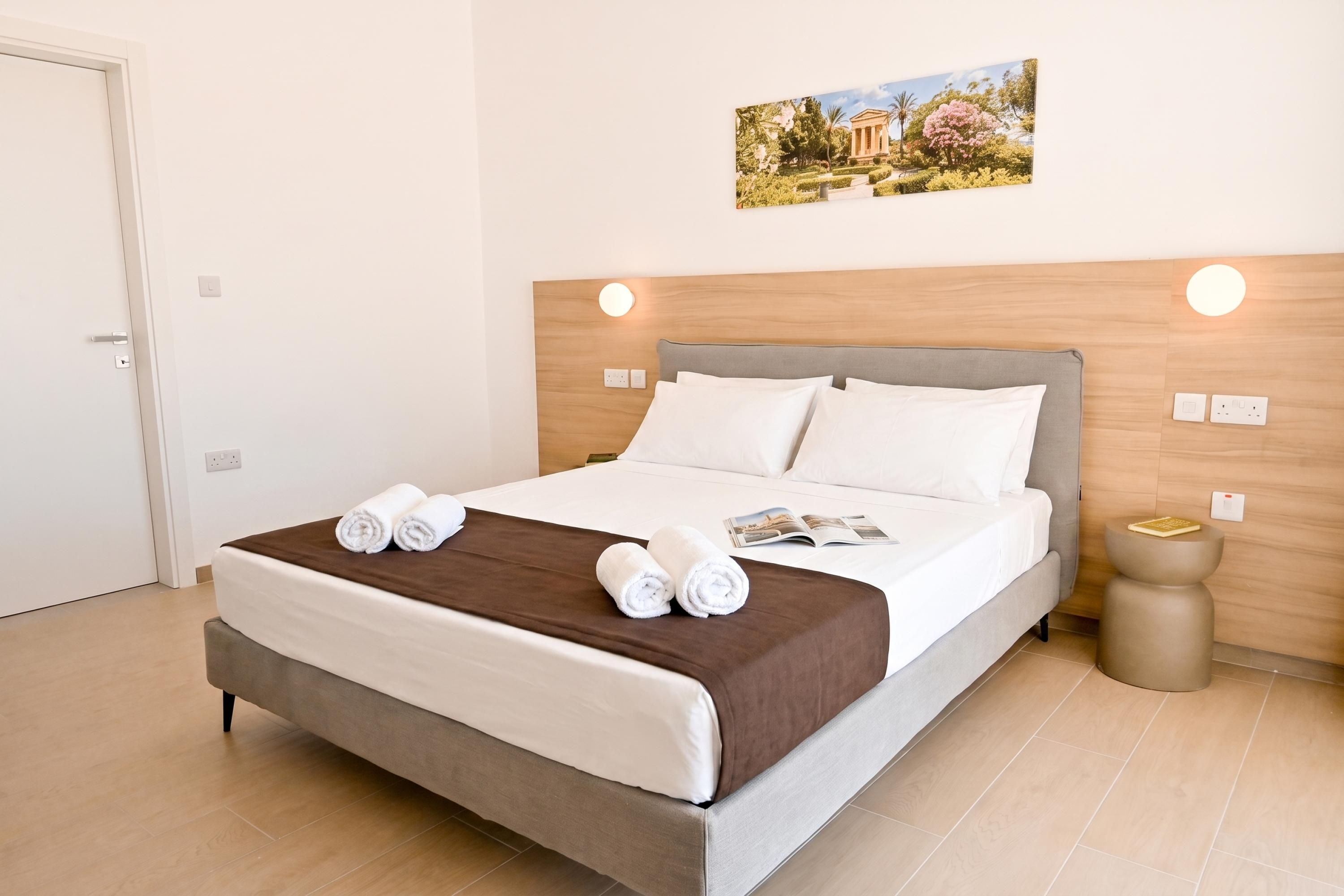Une chambre d’hôtel avec un lit, une table de chevet, une lampe fixée au mur et un tableau au-dessus du lit.