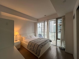 Un dormitorio con cama, mesita de noche, lámpara, ventana y vistas a edificios.