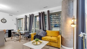 A modern living room with a yellow sofa, a dining table with chairs, a coffee table, and a large window with curtains. - StayToronto - The Entertainment Suites (Toronto)