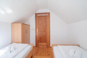 A small, well-lit room with two single beds, a wooden wardrobe, and a door leading to a lofted area.