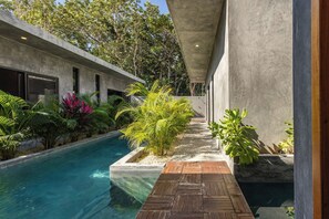 Un área de piscina moderna con cubierta de madera, rodeada de exuberante vegetación y una estructura de edificio de hormigón.
