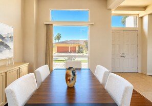 A conference room with a wooden table, white chairs, a vase, and a view of a house through a large window. - Palm Desert 4-Bedroom Retreat with a sparkling pool in the heart of Palm Desert (Palm Desert)