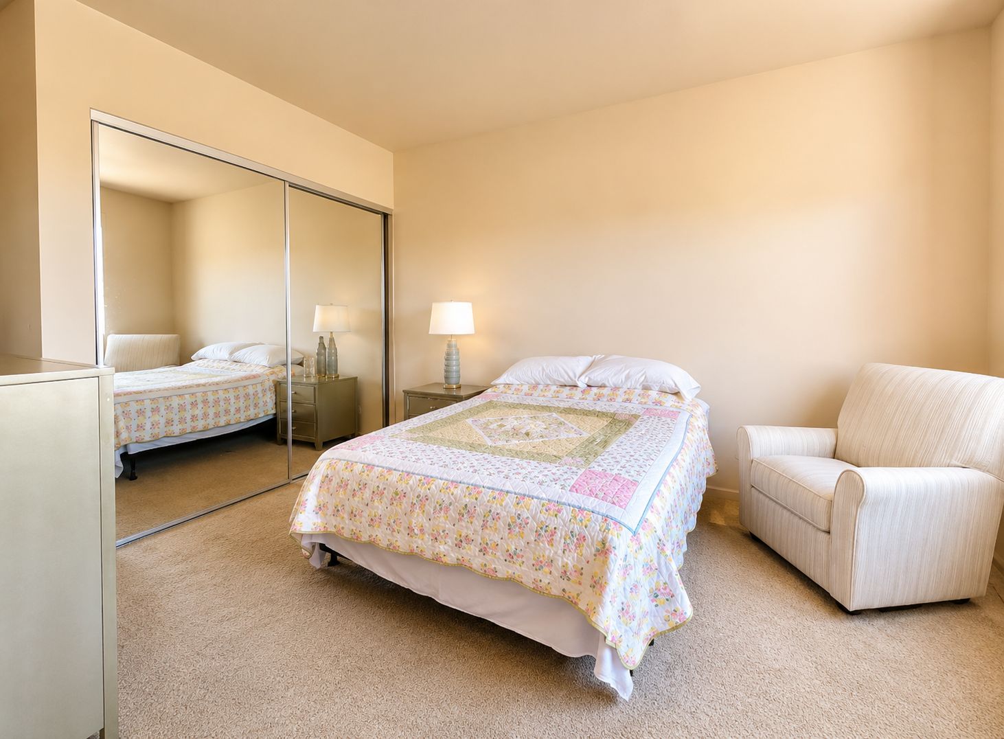 Une chambre à coucher avec un lit, une chaise, une table de chevet, une lampe et un miroir.
