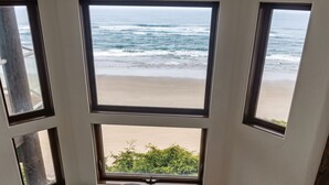 A room with a view of the beach through multiple windows, a wooden stool, and a potted plant. - Brewer’s Bluff | Iconic Ocean View Estate (Lincoln City)