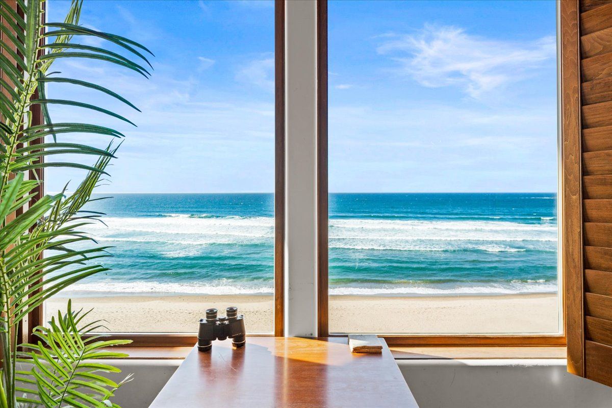 A room with a large window overlooking a beach, a wooden table, and binoculars.