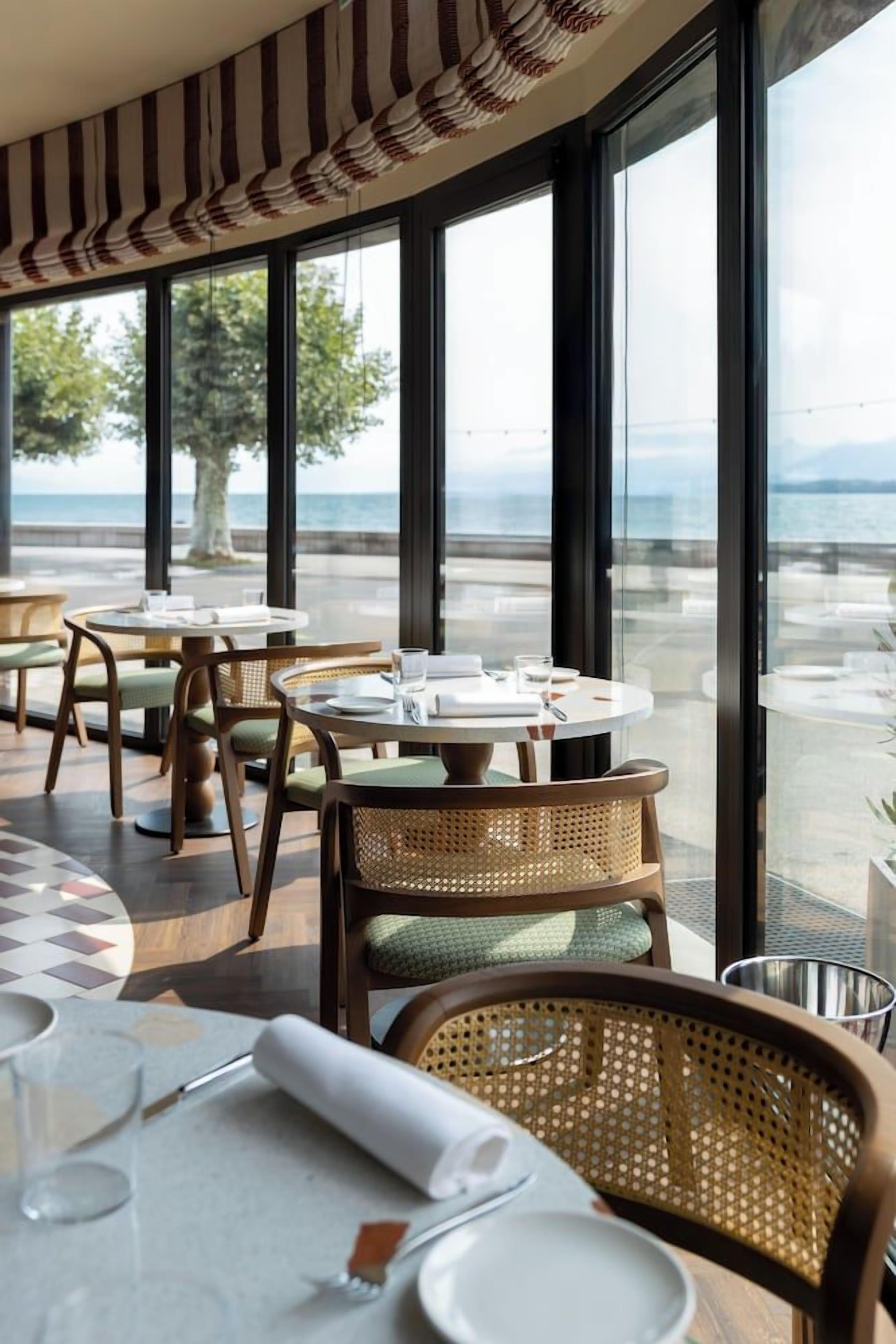 A restaurant with round tables and wicker chairs, large windows with a view of the sea.