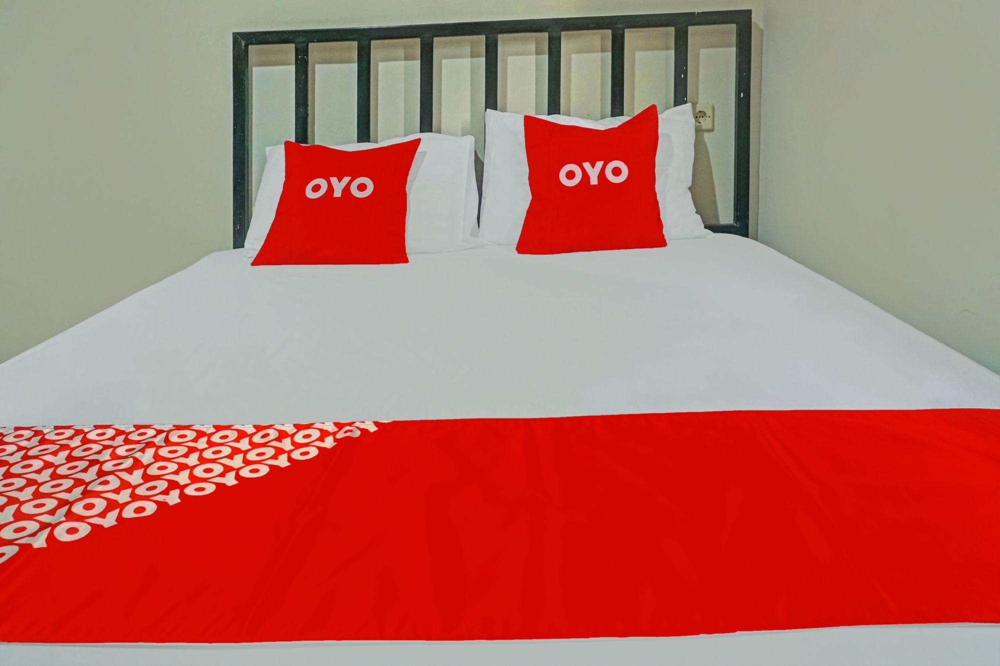 Ein Bett mit weißer und roter Bettdecke, zwei Kissen mit dem Schriftzug 'OYO' und ein schwarzes Metall-Kopfteil.